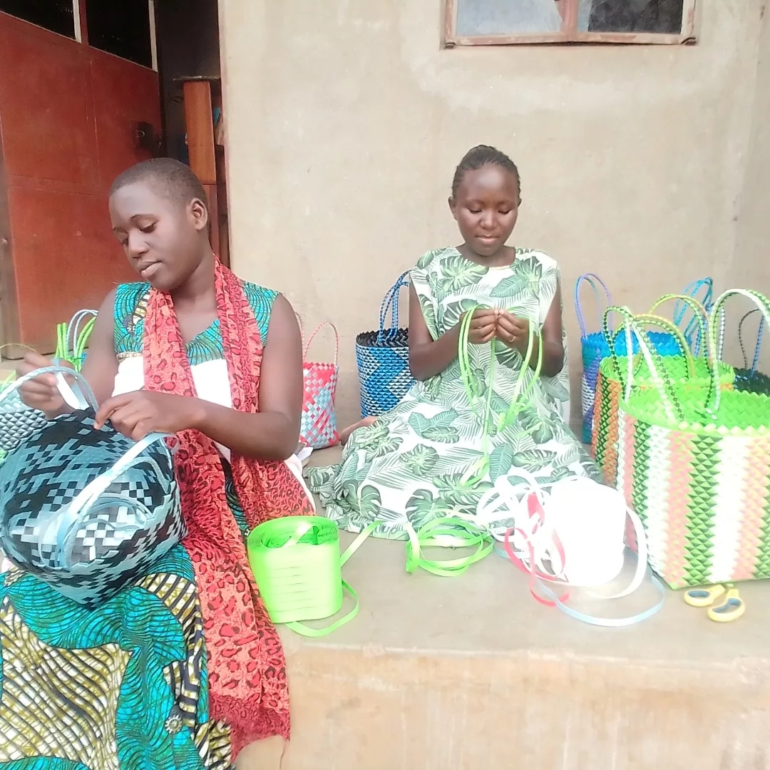 Two girls seated outside weaving handles onto colorful handmade bags, surrounded by finished woven baskets and crafting materials.