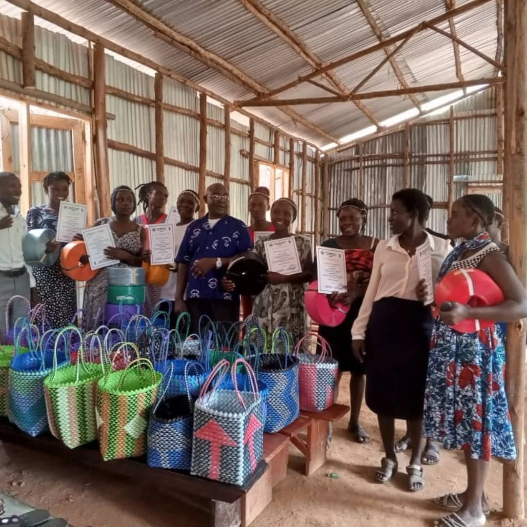 Group of women and girls holding certificates and handmade woven bags inside a corrugated metal community workshop building.