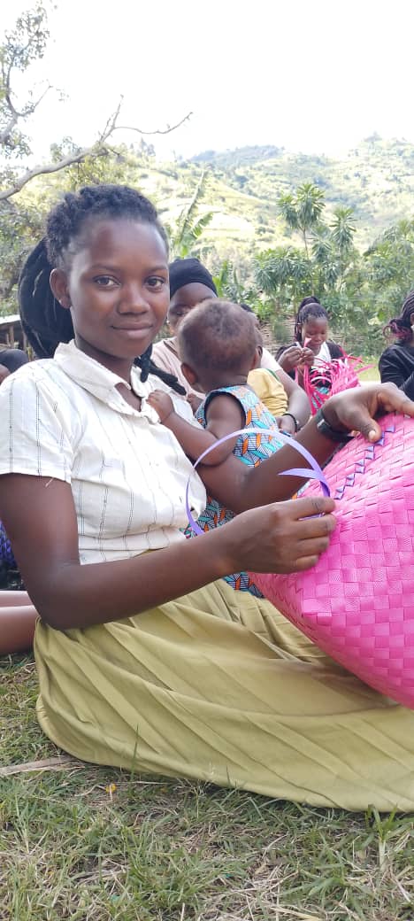 A young mother practices basket weaving with her baby beside her.